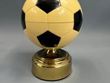 un ballon de football jaune et noir posé sur un socle doré, sur fond gris