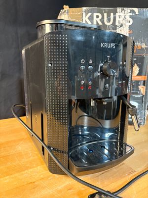 une cafetière posée sur une table en bois, à côté d'une boîte de Krups