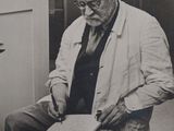 black and white photo of an old man sitting on a chair cutting a piece of paper