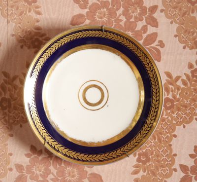 une assiette bleue et dorée, bordée d'or, posée sur un papier peint à motifs floraux