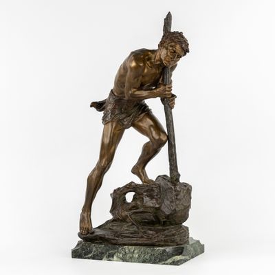une statue en bronze représentant un homme tenant une lance, assis sur un rocher