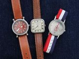 Trois montres sont posées sur une surface bleue, avec un bracelet rouge, blanc et bleu.