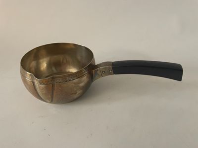 une vieille casserole en laiton avec un manche noir, posée sur une surface blanche