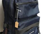 un sac à dos bleu muni d'une étiquette, posé contre un mur