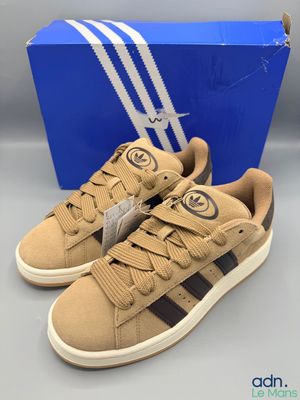 une paire de baskets Adidas de couleur beige et noire, accompagnée d'une boîte bleue