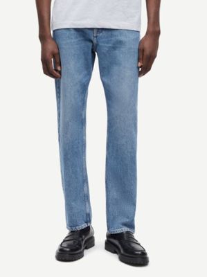 Un homme vêtu d'un t-shirt blanc et d'un jean se tient debout devant un fond blanc.