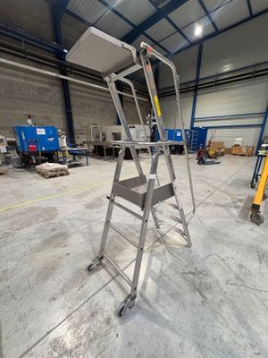 une photo représentant une échelle métallique dans un entrepôt, avec des cartons et des palettes en arrière-plan