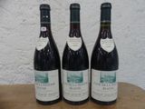 Trois bouteilles de vin rouge sont posées sur une table en bois devant un mur blanc.