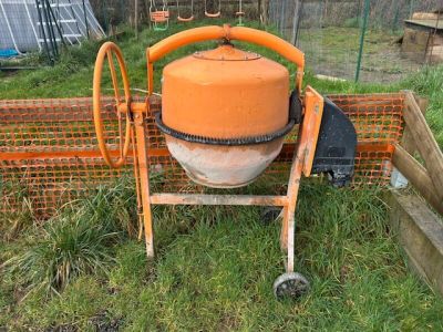 une bétonnière orange posée dans l'herbe, près d'une clôture