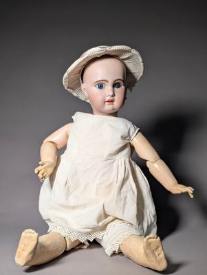 an old fashioned doll sitting on the ground wearing a white dress and a hat