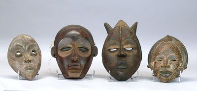 Quatre masques africains sont exposés sur des socles en acrylique posés sur une surface blanche.