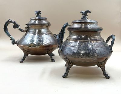 une paire de théières anciennes en métal argenté, posées sur un support, sur une table