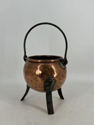 une image représentant un petit pot en cuivre muni d'une poignée, posé sur un support