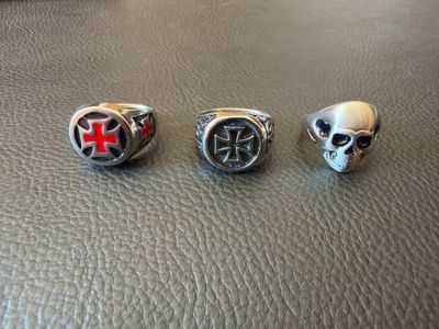 trois bagues en argent ornées de croix et de têtes de mort, posées sur une surface en cuir