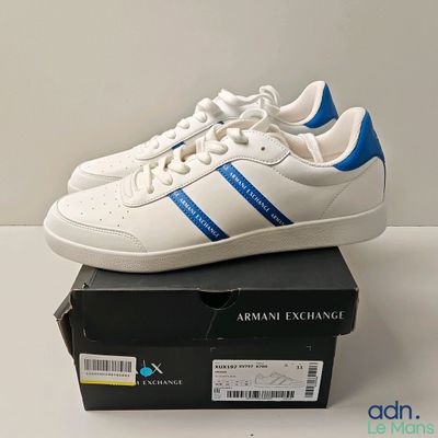 Baskets adidas Originals blanches et bleues, ornées de bandes bleues, posées sur une boîte
