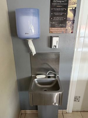 un lavabo en acier inoxydable dans des toilettes publiques, avec un sèche-mains bleu fixé au mur
