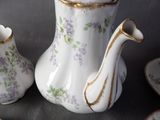 a white tea pot with purple flowers on it next to a plate and cups
