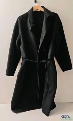 un manteau noir suspendu à un cintre sur un mur blanc, avec une chemise noire en dessous