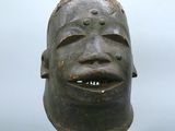 statue représentant le visage d'un homme posée sur un socle, avec un ciel nuageux en arrière-plan