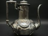 théière plaquée argent, ornée de motifs élaborés, posée sur un socle, sur fond noir