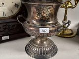 a silver vase on a table with a clock in the background