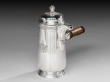 une cafetière ancienne en argent, dotée d'une poignée en bois, posée sur un support blanc