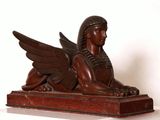 statue d'un sphinx ailé posée sur un socle en bois, avec un mur blanc en arrière-plan