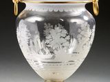 un vase en verre muni d'une anse dorée, posé sur un socle