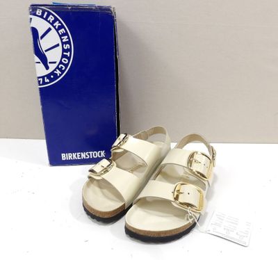 une paire de sandales Birkenstos blanches posées sur une surface blanche, à côté d'une boîte bleue