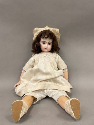 une poupée ancienne assise par terre, vêtue d'une robe blanche et d'un chapeau