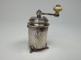 un moulin à café ancien en argent, muni d'un manche en bois, posé sur une surface blanche