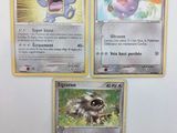 Trois cartes Pokémon sont posées sur une surface blanche bordée d'un liseré jaune.