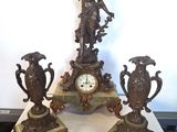 trois statues en bronze posées sur une table sur laquelle se trouve une horloge
