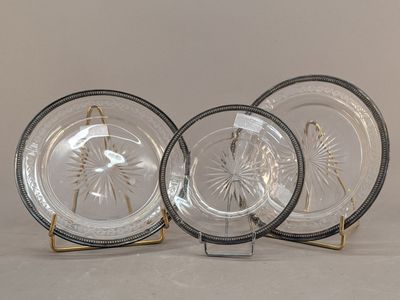 trois assiettes en verre transparent aux bords dorés, posées sur des socles, sur fond blanc