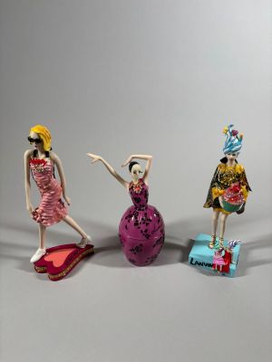 trois figurines représentant des femmes en robe et chapeau sont disposées devant un fond gris