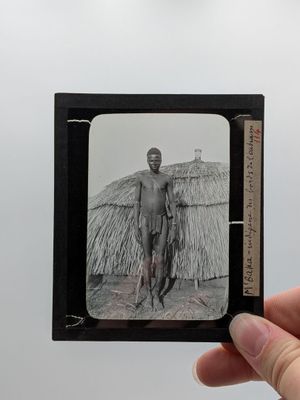 une main tenant une petite photo en noir et blanc représentant un homme devant une hutte au toit de chaume