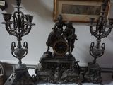 une horloge en argent posée sur une table, à côté de trois chandeliers