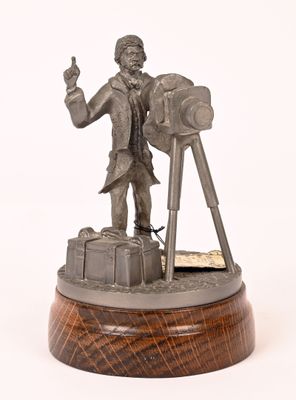 une statue représentant un homme muni d'un appareil photo, posée sur un socle en bois