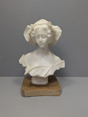 un buste blanc représentant une femme coiffée d'un chapeau, posé sur un socle en bois