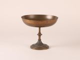 an antique brass footed bowl on a pedestal on a white background