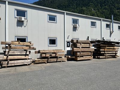 Plusieurs piles de bois sont empilées devant un bâtiment, avec une montagne en arrière-plan.