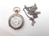 une montre de poche en argent et une chaîne posées sur une surface blanche