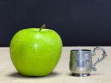 a green apple next to a silver cup on a table