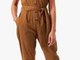 a woman wearing a brown jumpsuit and sandals stands in front of a white background