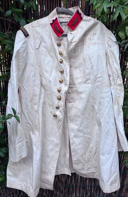 une veste beige à col et boutons rouges, suspendue à un cintre devant une clôture en bambou