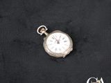 une image représentant une montre de poche en argent posée sur un tissu noir