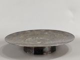 une assiette en argent posée sur un socle noir, sur une surface blanche