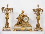 une horloge en or posée sur une table, à côté de trois chandeliers