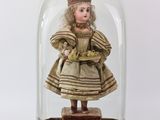 une image représentant une poupée miniature placée dans une cloche en verre posée sur un socle en bois