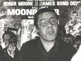 photographie en noir et blanc d'un homme à lunettes debout devant une affiche de cinéma